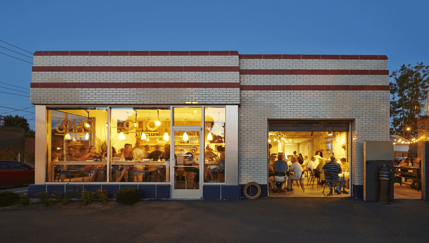 Modern restaurant in a converted brick building with diners visible through a large window and an open garage door at dusk